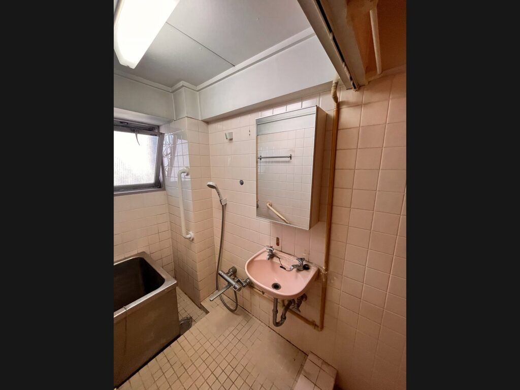 リフォーム前の浴室内洗面。タイル壁に古いピンクの壁付け洗面台と露出配管、浴槽と同じ空間に設置